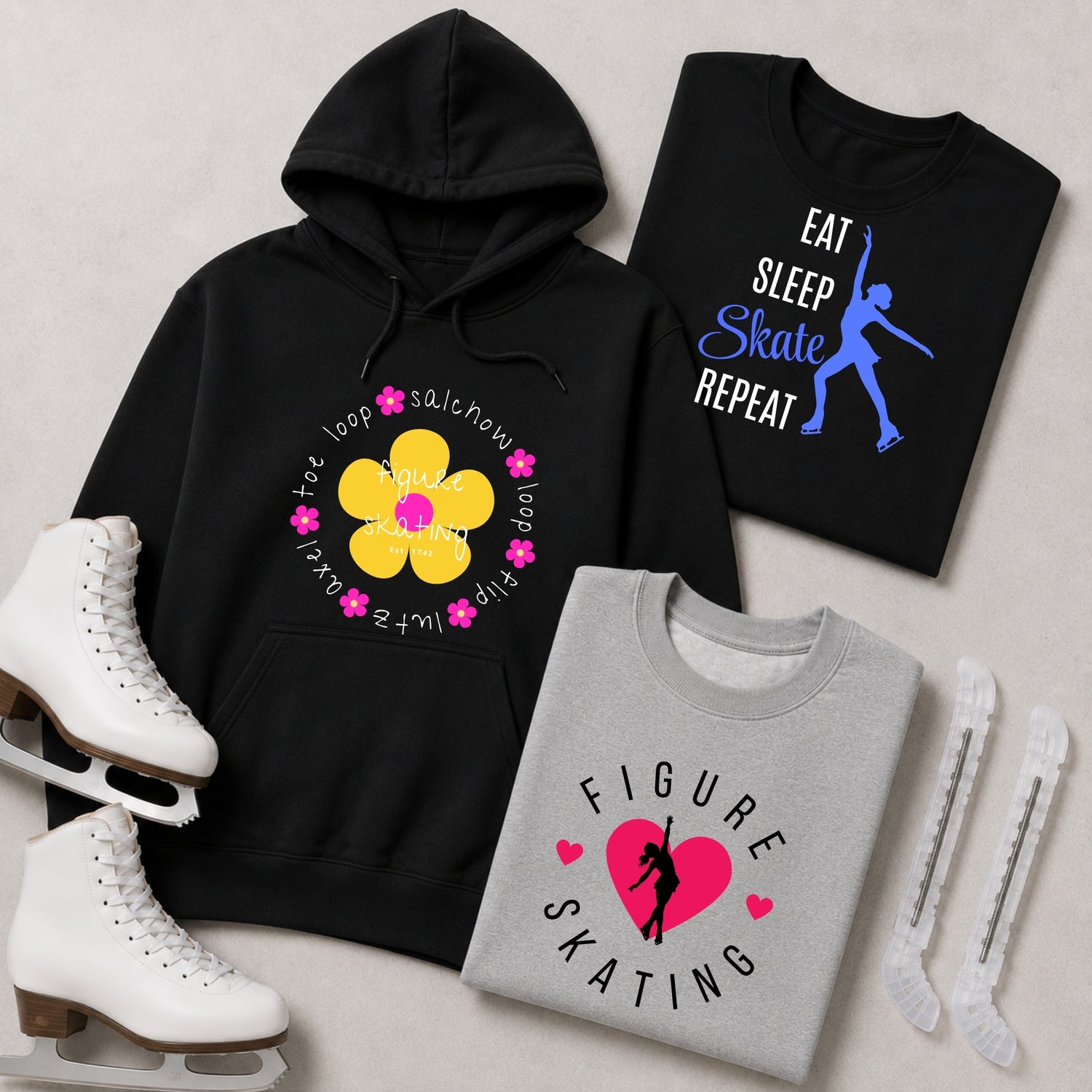 Three graphic sweatshirts and ice skates arranged on a light gray surface. The black hoodie has a yellow flower graphic with pink text reading 'toe loop flip and other moves'.