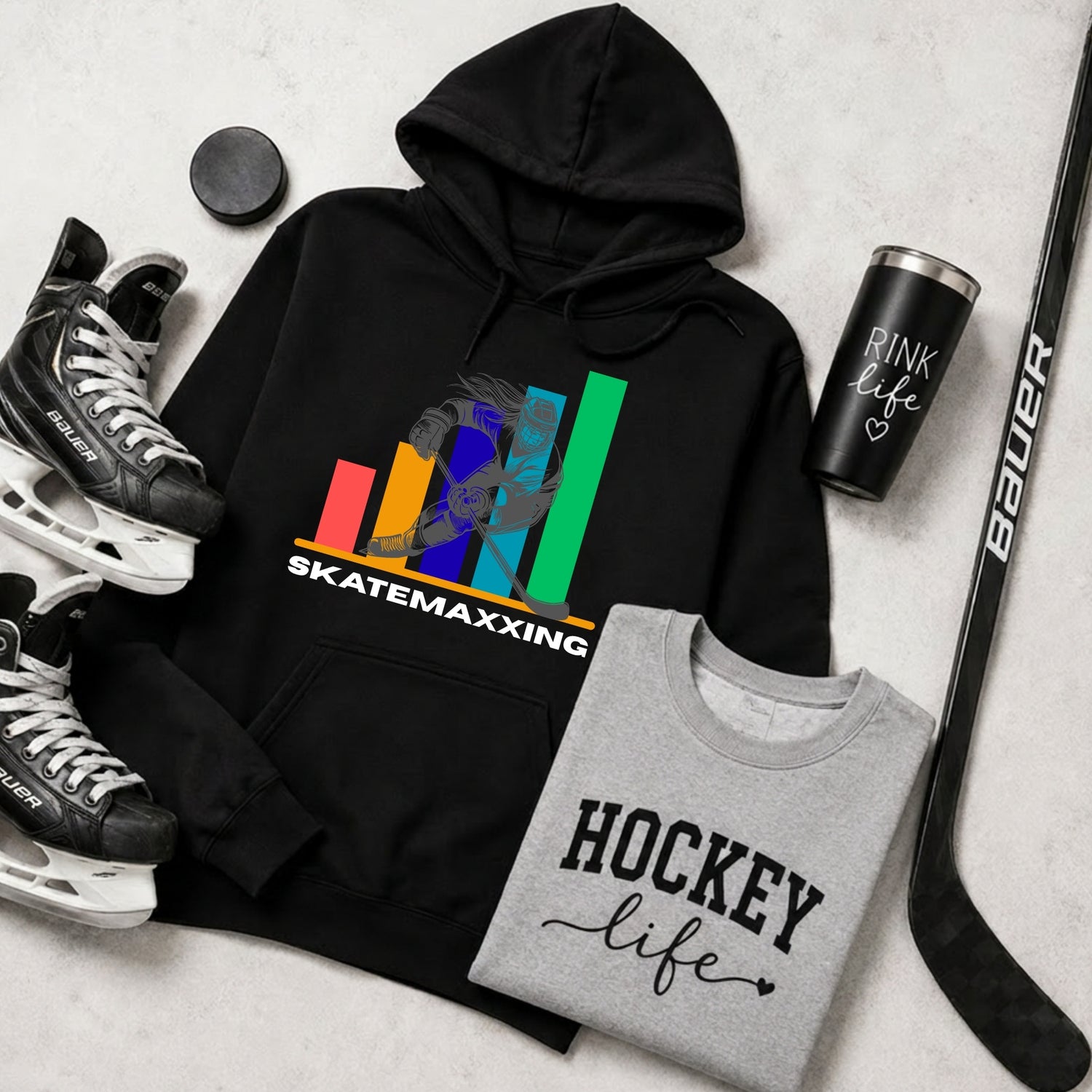 A flat lay of hockey-themed tops: a black hoodie with a colorful skater silhouette and the text 'SKATEMAXXING,' a heather gray T-shirt with 'HOCKEY life' printed in black, a black travel tumbler reading 'RINK life,' hockey skates, a hockey stick, and a black puck.