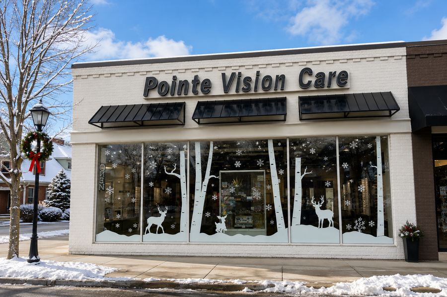 Front exterior of a storefront building labeled 'Pointe Vision Care' with large windows, black awnings, snow on the ground, bare trees, and holiday window decorations including white deer silhouettes and snowflakes.