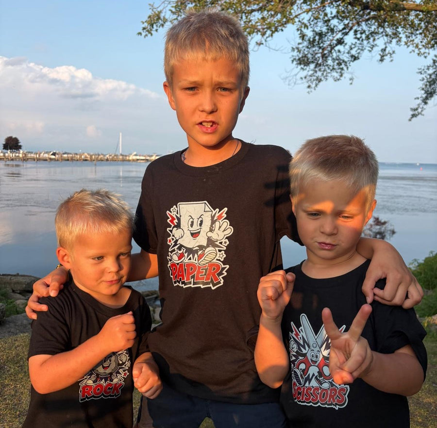 Three boys standing outdoors by the water wearing black graphic T-shirts. The center boy wears a shirt with a cartoon character and the word 'Paper', the left boy wears a shirt with 'Rock' and a skull graphic, and the right boy wears a shirt with 'Scissors' and a robot graphic.